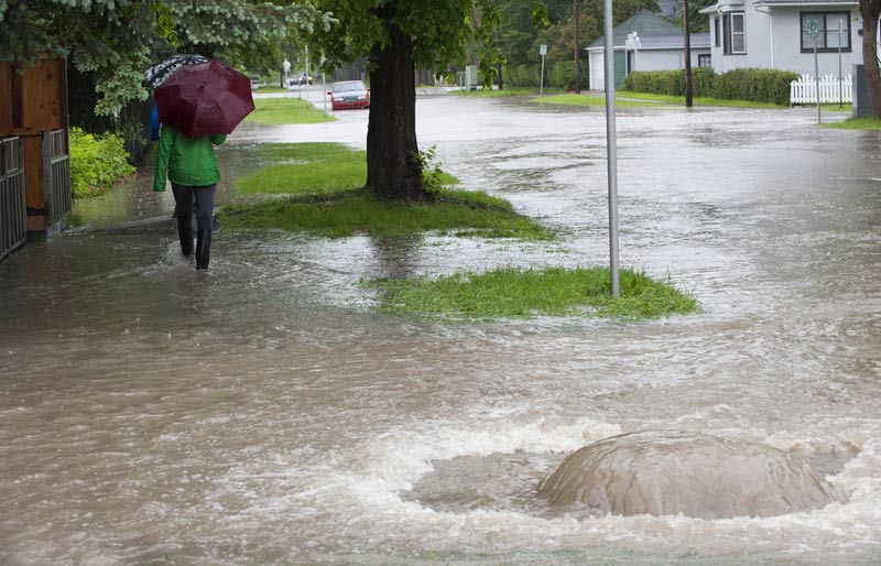 Sous la pluie, une personne munie d’un parapluie passe devant un flot d’eau trouble qui jaillit d’une bouche d’égout.