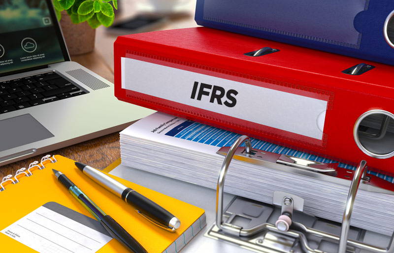 A close-up of a desk with pens, pads, a laptop and a number of binders one of which is labeled IFRS. 