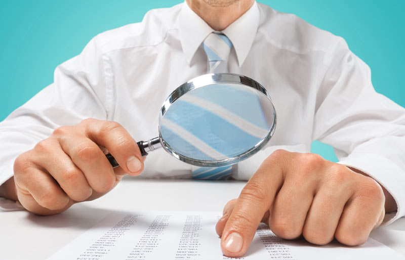 This is a photograph of a business professional looking at a spread sheet through a magnifying glass. 