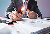 Business person using a magnifying glass to look at large book of business documents. 