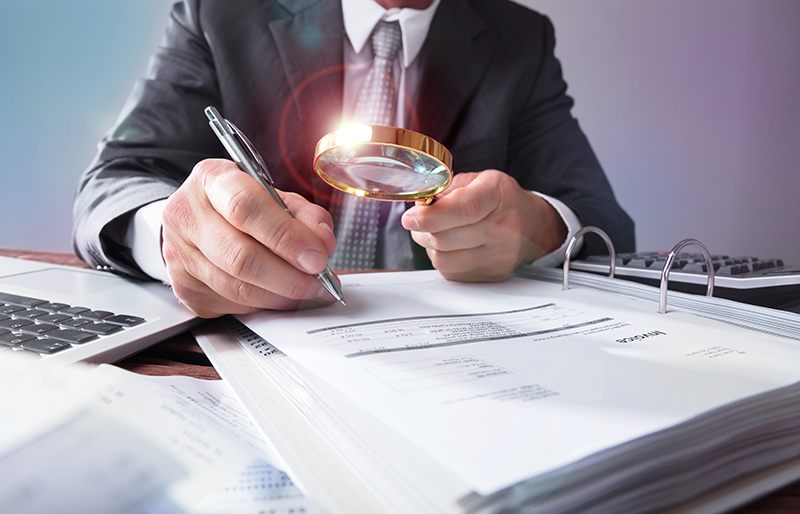 Business person using a magnifying glass to look at large book of business documents. 