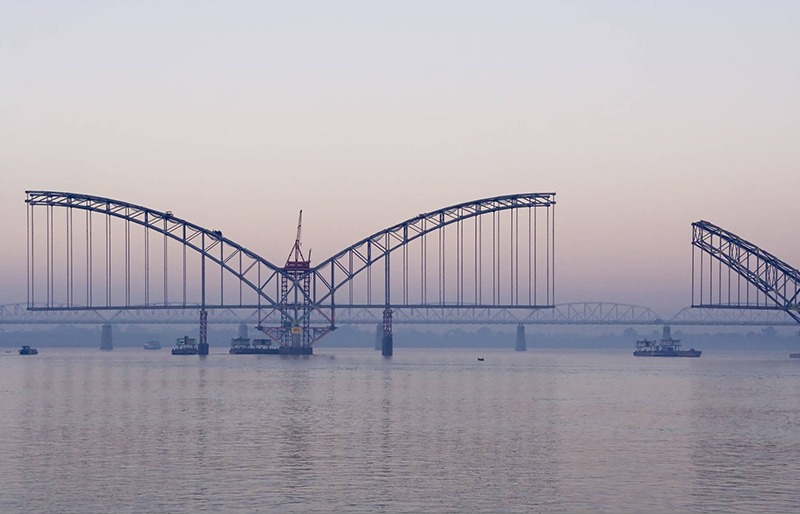 Cadrage d'un pont en construction au-dessus de l'eau, un second pont et des bateaux sont visibles en fond.