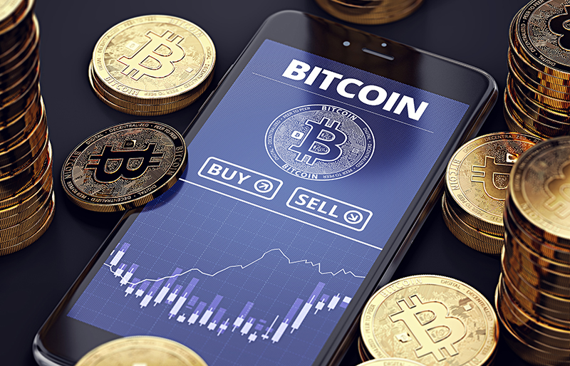 Une application de marché Bitcoin est affichée sur un cellulaire posé sur une table, ce dernier entouré de piles de monnaie Bitcoin.