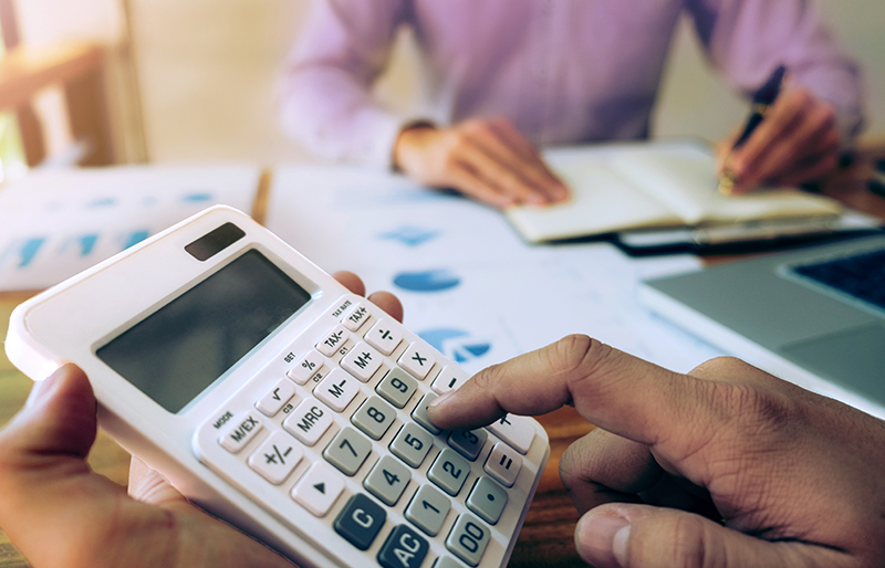 Cadrage sur les mains d’un comptable qui utilise une calculatrice et en fond, un professionnel derrière une table prend des notes, des documents financiers posés devant lui.