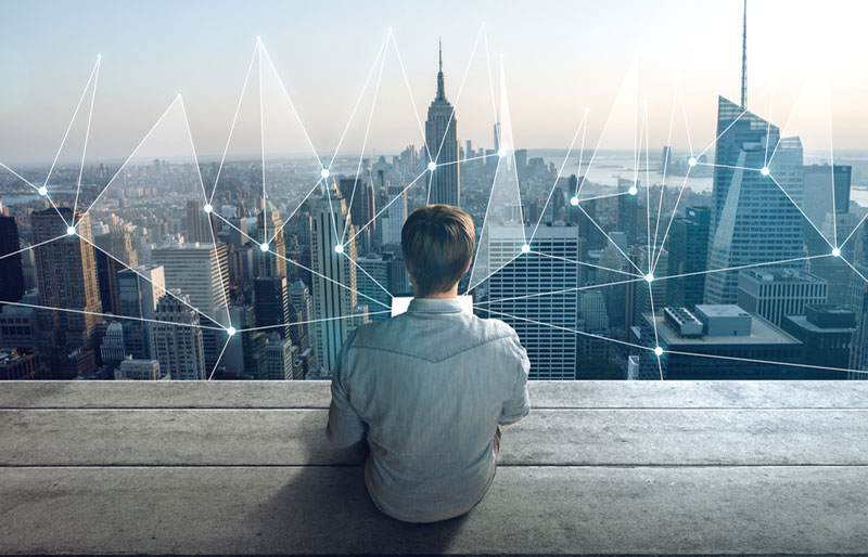 Man with laptop looking at cityscape with overlay of lines connecting various buildings together.