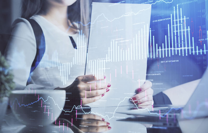 A businesswoman sitting at a desk reading papers, superimposed with graphs.