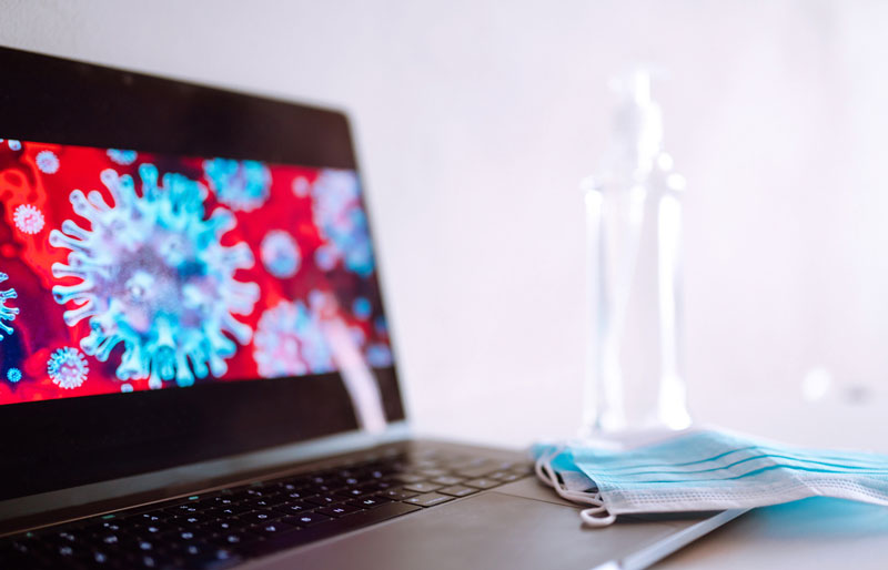 Desktop with mask, hand sanitizer and laptop showing microscopic image of virus.