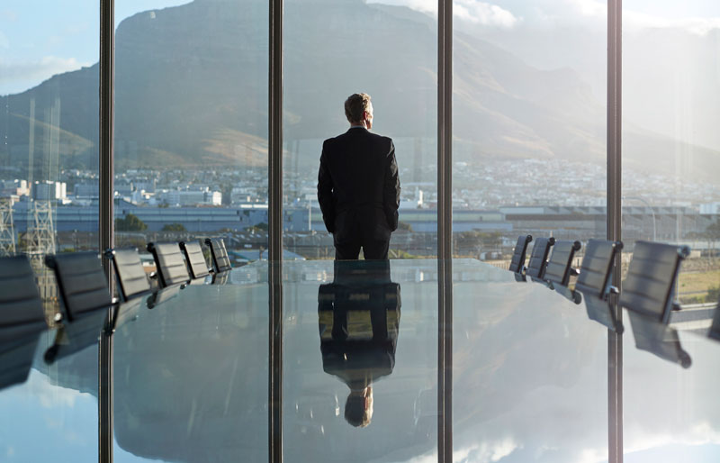 Businessperson looks out panoramic office window, view of mountains is reflected in large boardroom table.
