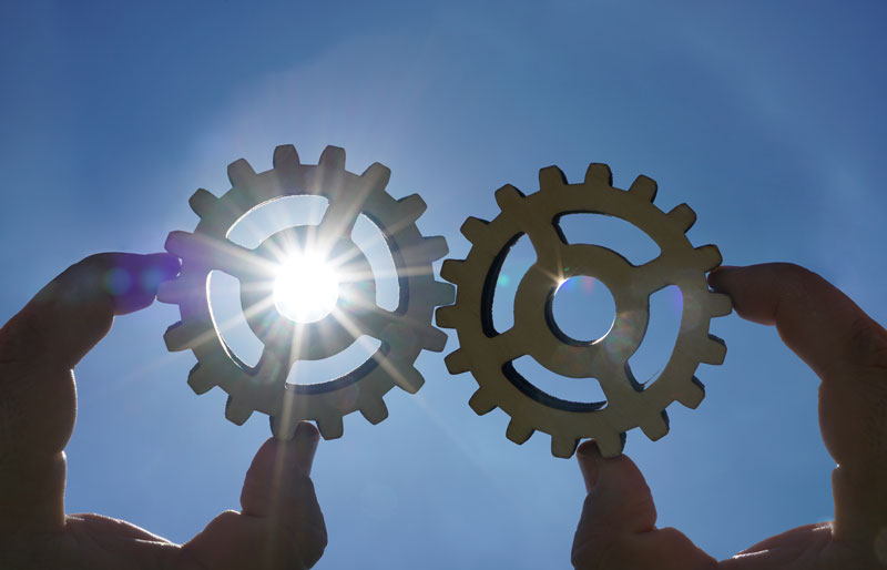 Two hands, each holding a cogwheel, sun and blue sky in background.