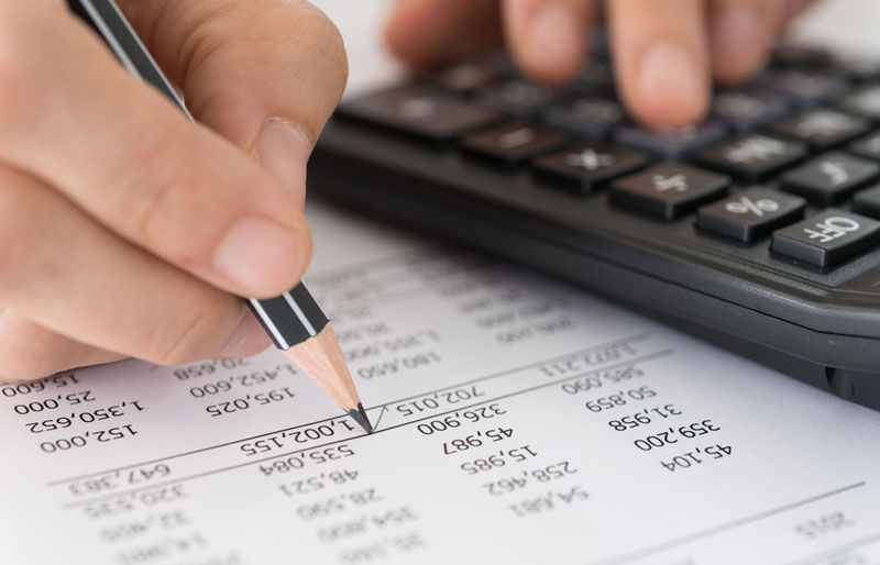 L’analyse de données en audit est illustrée par un gros plan sur les mains d’une personne qui examine des colonnes de chiffres à l’aide d’un crayon et d’une calculatrice.