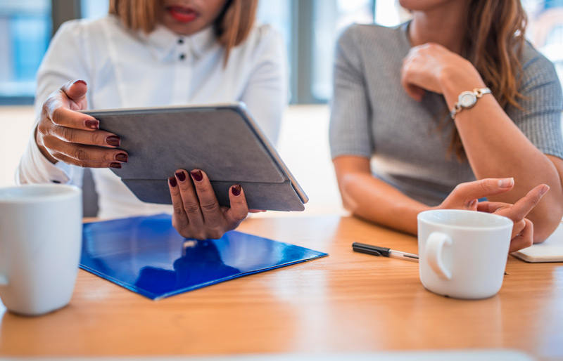 Two businesswomen look at a tablet while sitting at a desk.