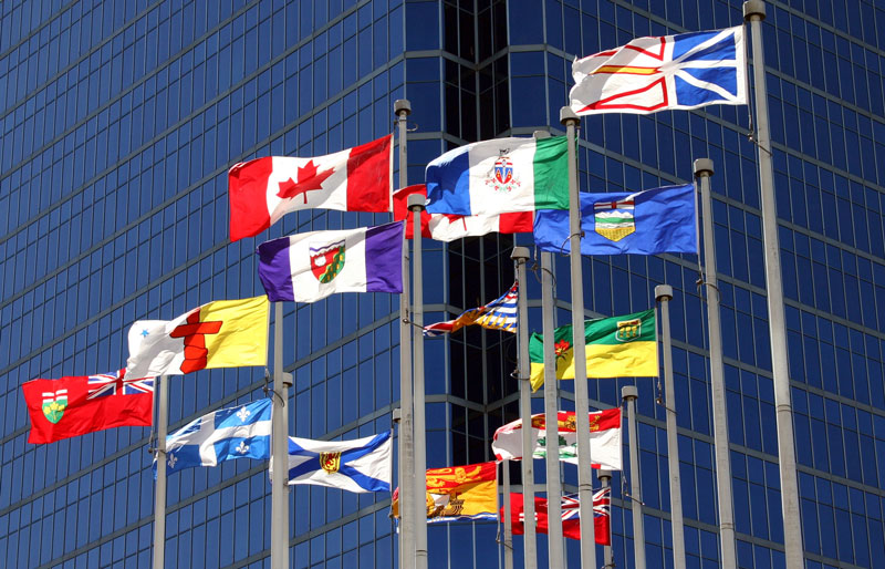 Flags for Canada and each of the provinces and territories on flagpoles in front of a high rise.