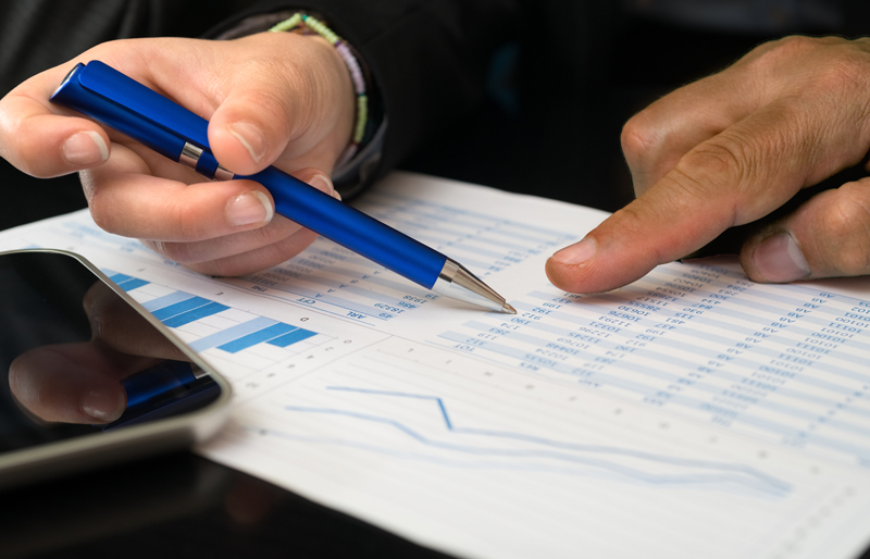 Gros plan sur les mains d'un professionnel qui consulte des documents financiers, stylo bleu à la main.