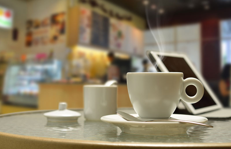 A photograph of a coffee shop table with a coffee, tablet PC and sugar bowl. 