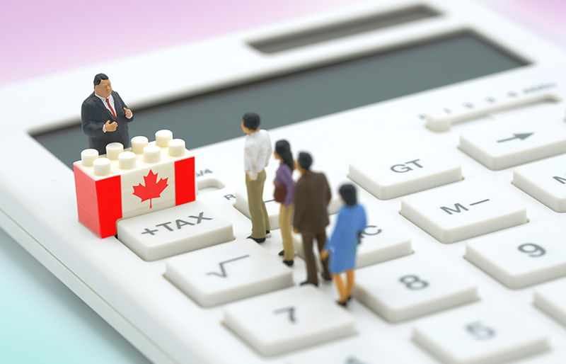 Miniature toy people lined-up to a toy building-block with the Canadian flag and a miniature toy business person behind, upon a calculator  