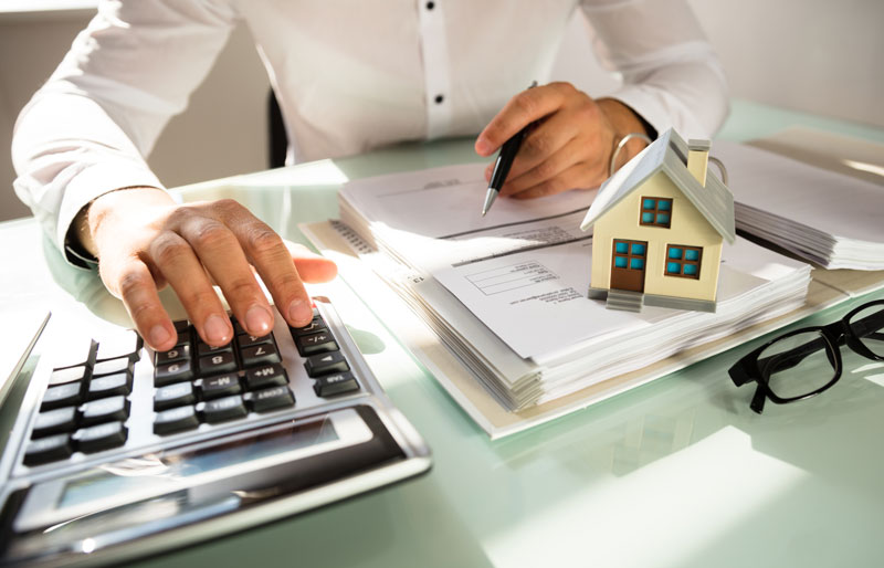 Businessperson using calculator at desk with papers and small house model.