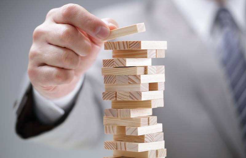 Un homme d’affaires pose prudemment une pièce de jeu de construction en bois au sommet d’une tour composée de multiples autres pièces en bois.